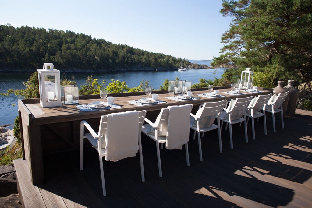 Langt trebord med hvite stoler og borddekking på terrasse ved blå fjord og grønn skog