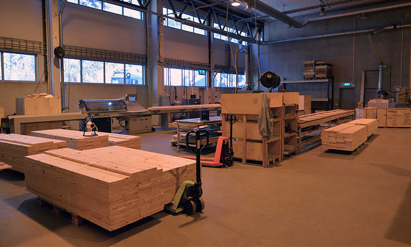 Stor industribygg med store stabler av lyse treplanker på grått gulv og palletruck.