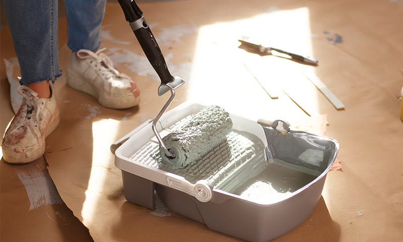 Grå malerulle i plasttraug med lys grå maling på papp, malersko i bakgrunnen