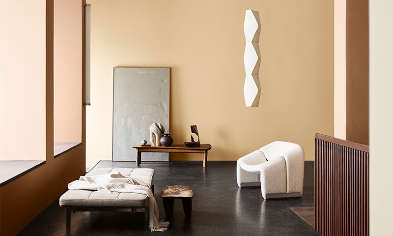 Minimalistisk beige stue med mørkt gulv, lav benk, hvit lenestol, speil, trebord og hengelampe.