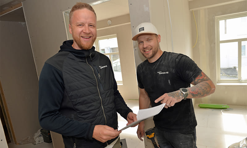 To håndverkere i svarte arbeidsklær diskuterer papirer i en lys, uferdig stue.