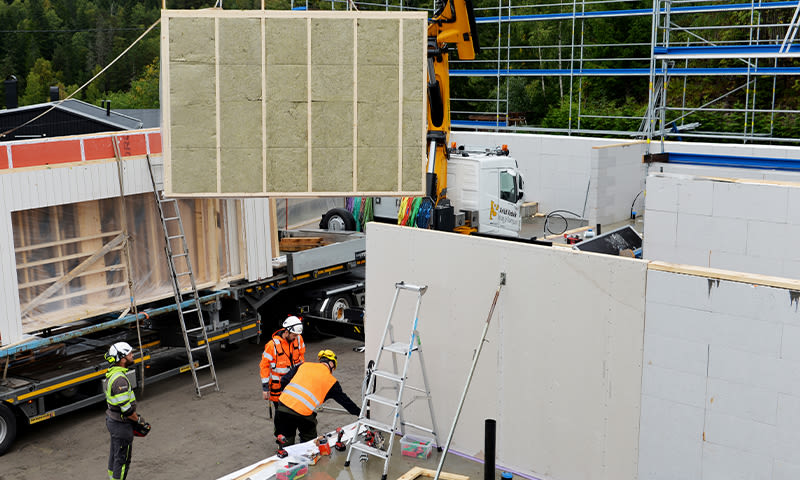 Gul kran løfter stor isolasjonsvegg, arbeidere i oransje vester ved uferdig hus