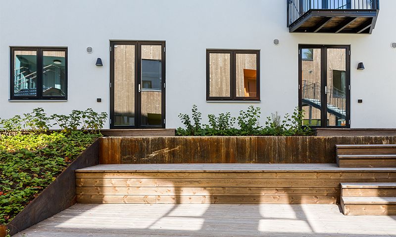 Hvit fasade med svartinnrammede glassdører og vinduer, tretrapp, balkong og grønn beplantning.