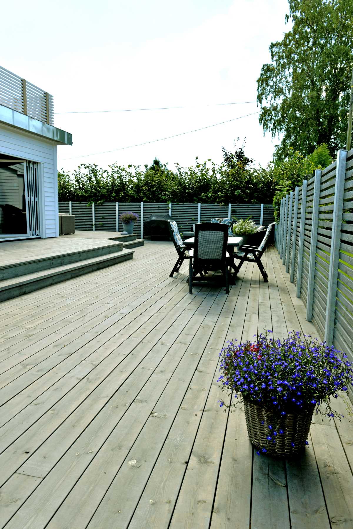 Stor treterrasse med grå gjerder, utebord med stoler og kurv med lilla blomster