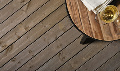 Delvis synlig rundt trebord med hvitvinsglass og serviett på mørkbrunt terrassegulv.