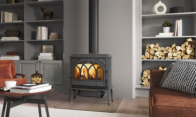 Svart vedovn med flammer, grå bokhyller med ved, brun sofa og trebord