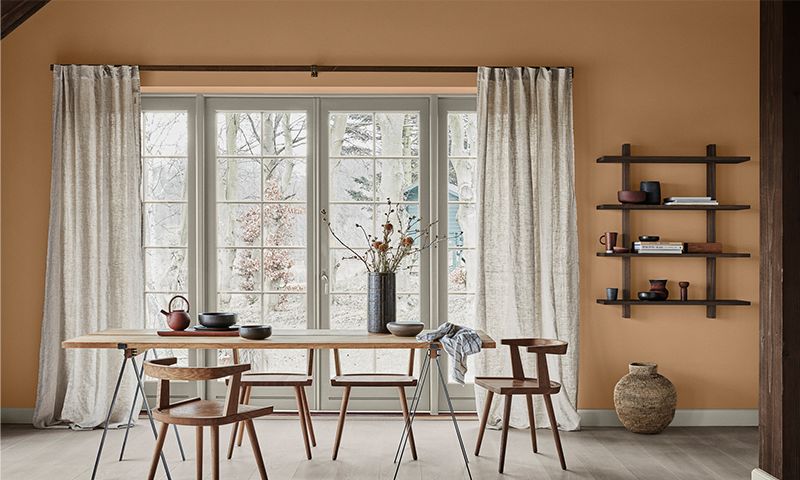 Spisebord med trestoler foran vindu, brun-oransje vegg, keramikk og vase med tørkede kvister.