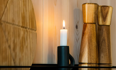 Tent hvitt stearinlys i svart lysestake på hylle med rund trefjøl og to høye tre-krydderkverner