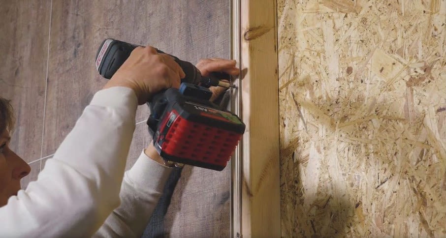 Person bruker svart-rød batteridrill for å feste trelist på vegg med lyse OSB-plater.