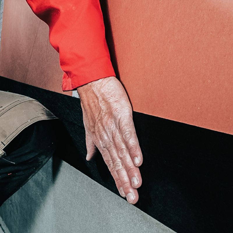 Hånd i rødt erme stryker svart matte mot grå gulvplate foran oransje vegg.