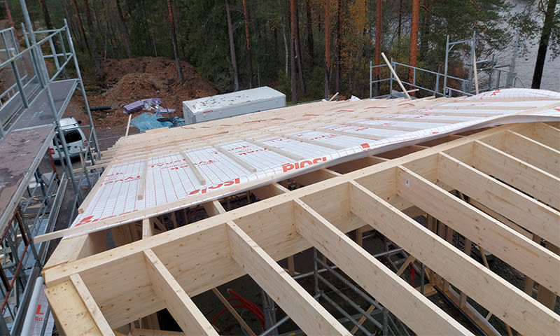 Uferdig tak med lyse trekonstruksjoner og hvit isolasjonsduk med rødt trykk, skog og innsjø.