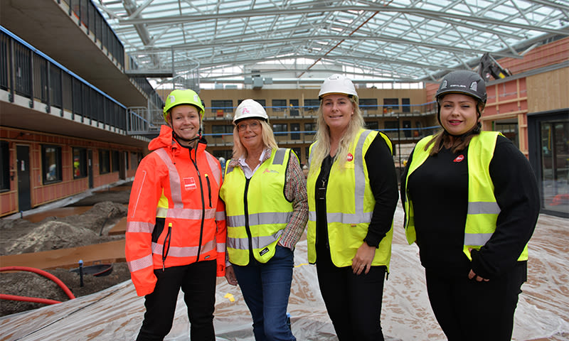 Fire personer i gule og oransje vernevester og hjelmer inne i uferdig bygg med glasstak