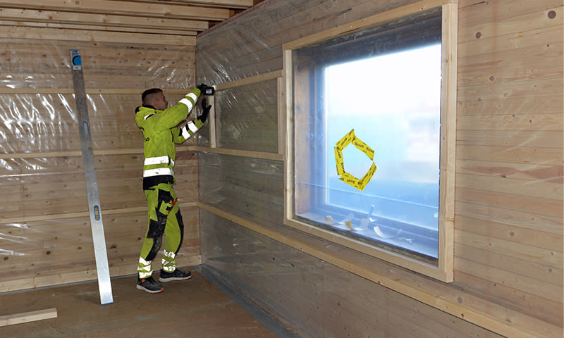 Arbeider i grønn refleksdress fester plastfolie på innvendig trepanel ved vindu.