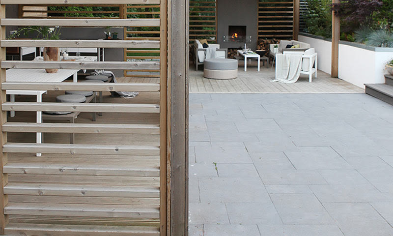 Moderne uteplass med grå steinfliser, pergola av lyst tre, hvite møbler og levende ild i peis