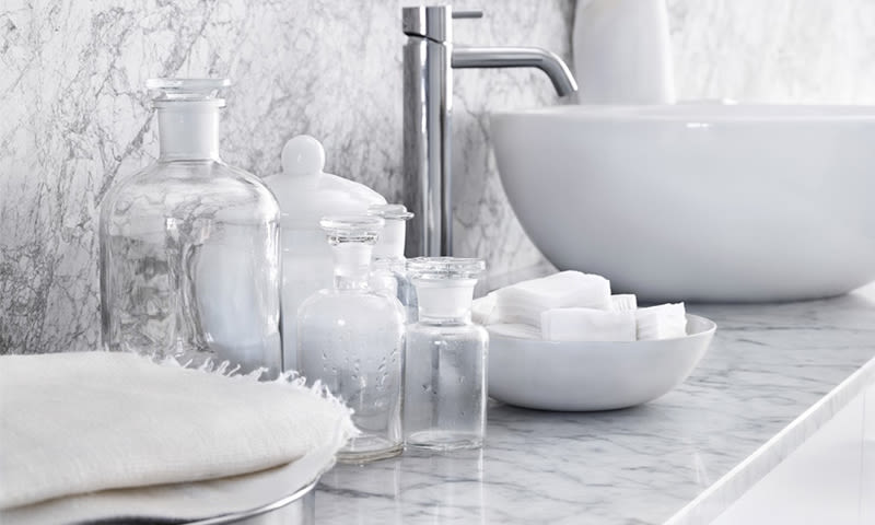 Baderomsbenk i hvit marmor med vask, krom armatur, glassflasker og bomullspads.