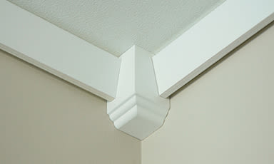 Hvitt taklisthjørne med dekorativ midtblokk mot beige vegg.