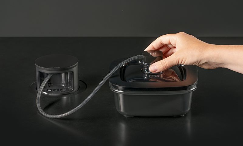 Hånd løfter gjennomsiktig beholder med grå slange fra svart innfelt kjøkkenavtrekk på svart benk