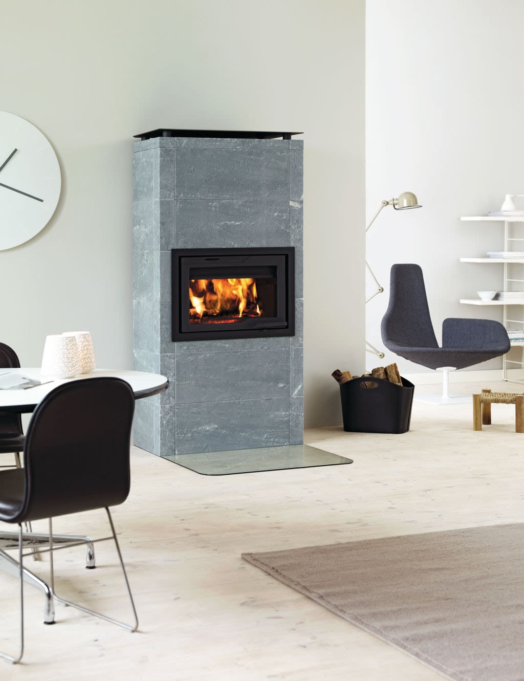 Moderne lyst rom med grå klebersteinspeis med flamme, vedkurv, hvit veggur og grå lenestol