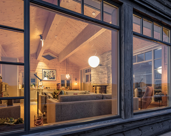 Utsikt gjennom vindu til lys trehytte med steinpeis, grå sofa, pendellampe og person.