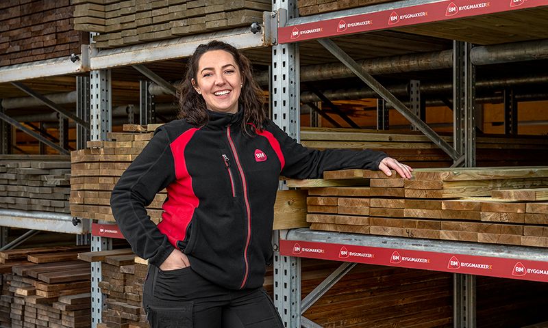 Ansatt i svart-rødt arbeidstøy står ved stabler med treplanker i lagerhall.