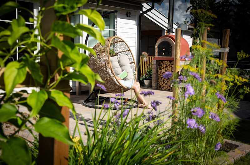 Person leser i kurvformet hengestol på lys terrasse med lilla blomster og vedovn.