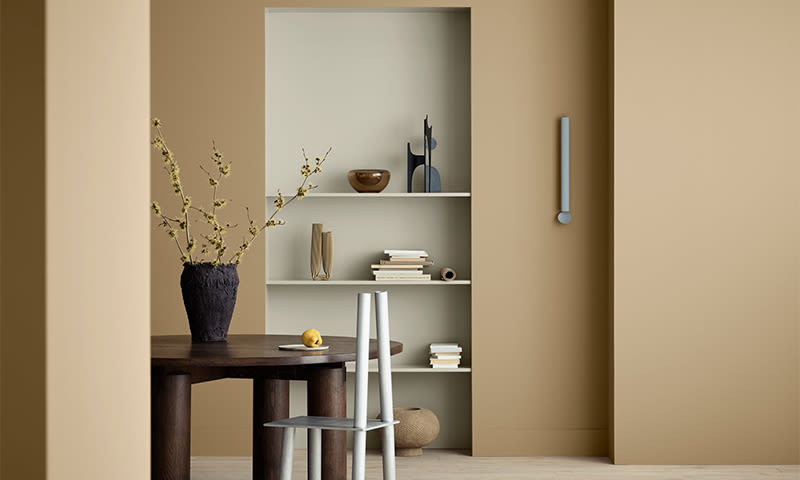 Beige rom med innebygde hyller, mørkt trebord, hvit stol og vase med gule blomster.