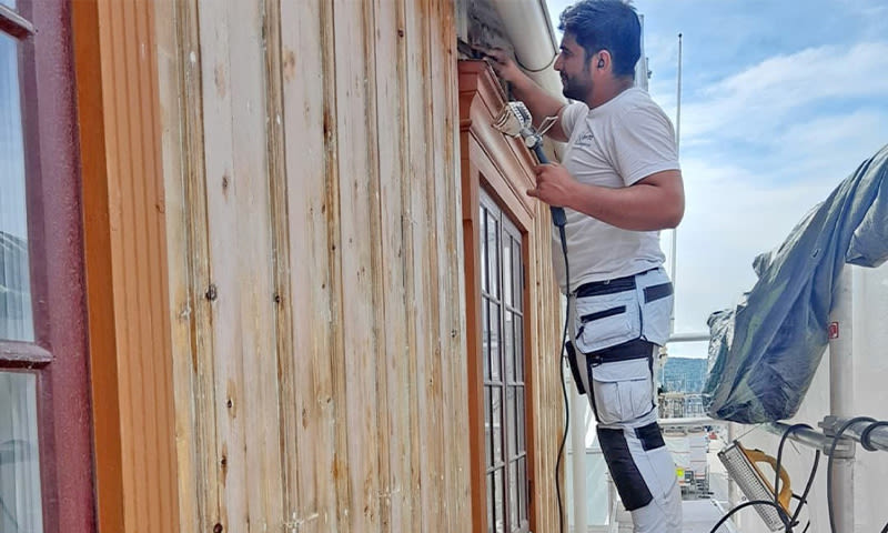 Håndverker på stillas bruker drill på oransjebrunt trepanel på husfasade mot blå himmel