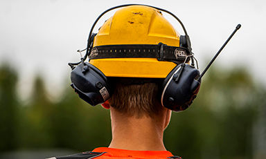Mann bakfra med gul hjelm, svart hørselsvern og oransje vest foran uskarp skog