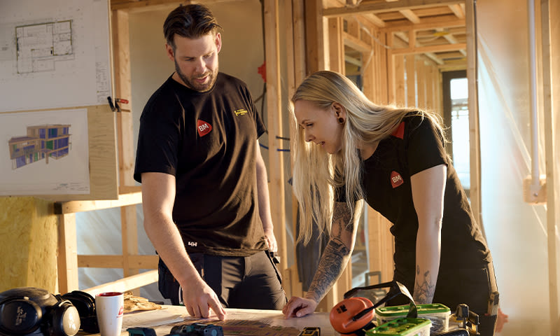 To håndverkere i svarte BM-skjorter studerer byggeplaner på trebord i råbygg med verktøy.