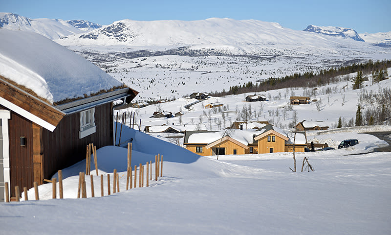 Oversiktsbilde av gule og brune hytter dekket av snø i solfylt fjellandskap med blå himmel