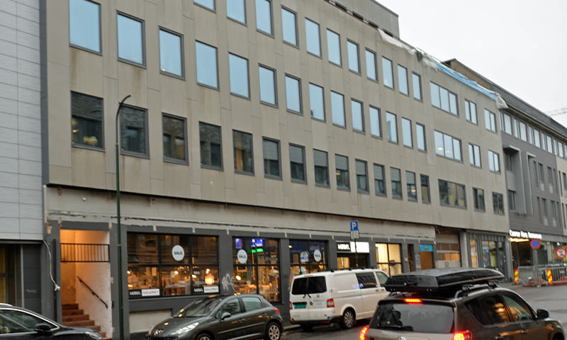 Beige fleretasjes kontorbygg med butikkvinduer, parkert hvit varebil og småbiler under grå himmel