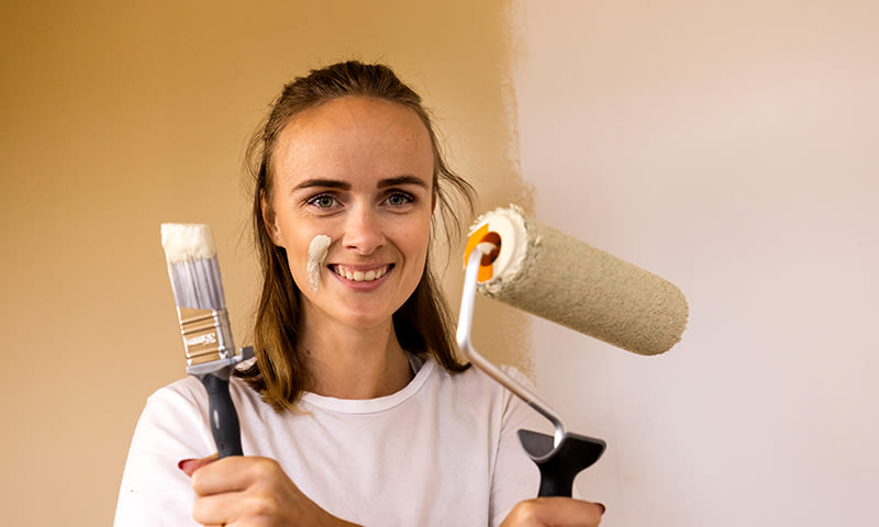 Person i hvit t-skjorte maler vegg i lyse beige toner med pensel og malerull