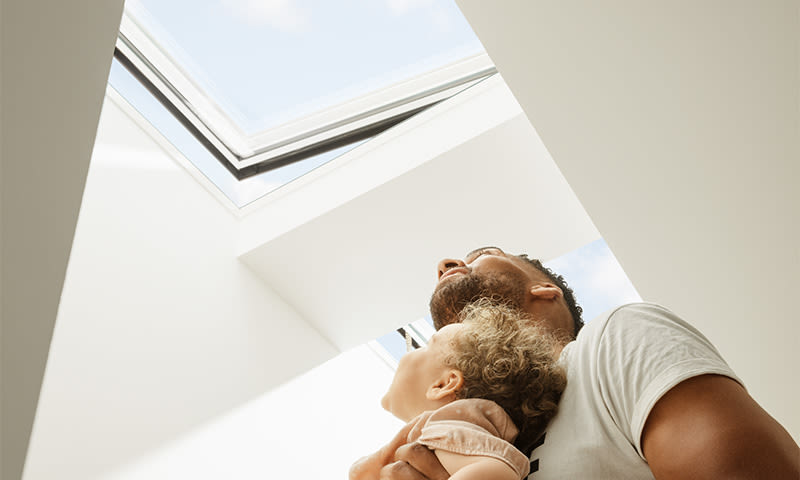 Far med skjegg løfter barn under takluke i hvitt rom mot blå himmel