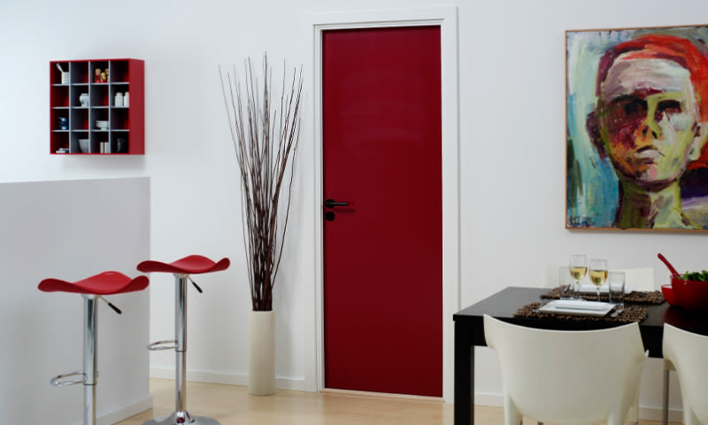 Hvitt interiør med rød dør, røde barkrakker, høy kvistvase, svart spisebord og hvite stoler.