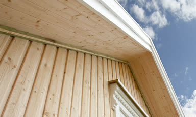 Skarpt vinkelbilde av lyst trekledning på hus med hvit taklist mot blå himmel