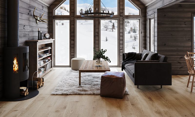 Rustikk tømmerstue med peis, mørk sofa, trebord og snødekt fjellutsikt gjennom høye vinduer