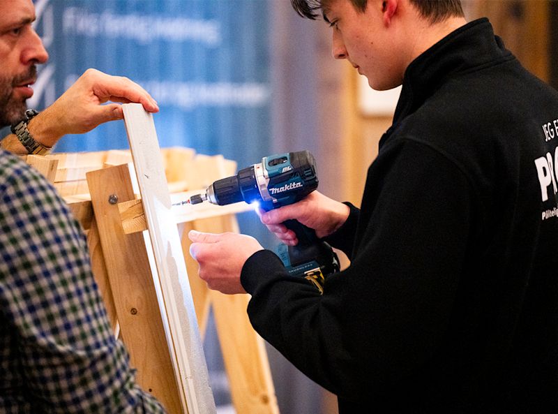 Ung person i svart genser bruker en Makita-drill på en trekonstruksjon, veiledet av en voksen mann.