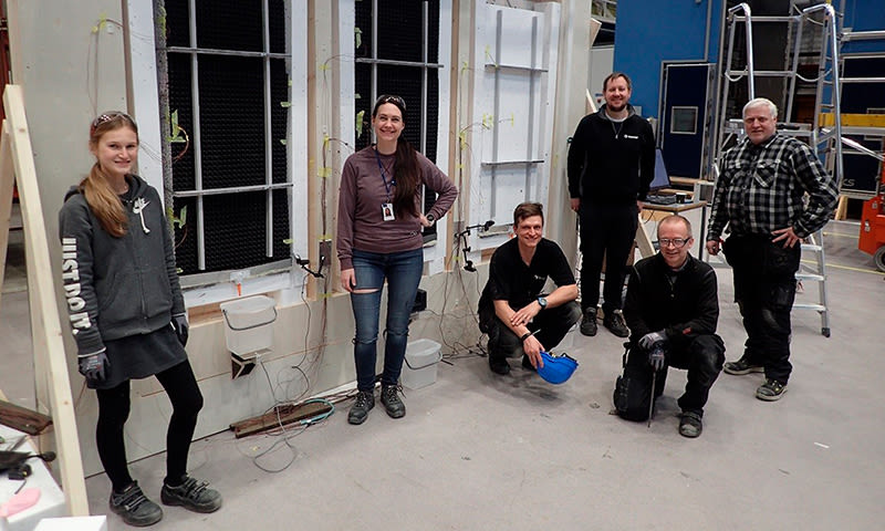Gruppe i verksted foran hvit vegg med kabler, bøtter og verktøy, grått gulv og blå bakgrunn
