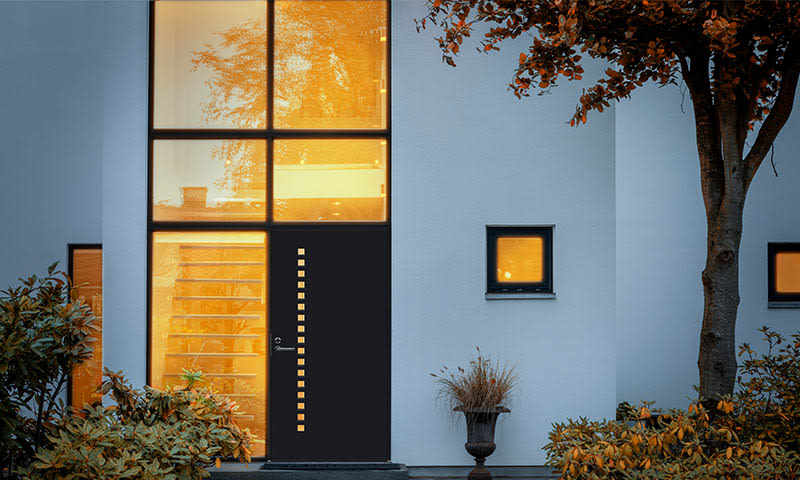 Hvit fasade med opplyst stort vindu, svart dør med små vindusglugger og høstgult løv.