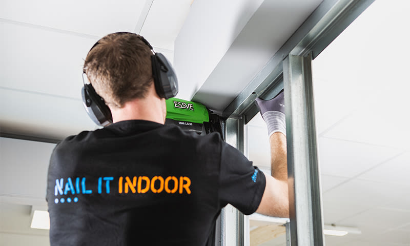 Mann bruker grønn ESSVE-spikerpistol på metallstender, iført hørselsvern og NAIL IT INDOOR t-skjorte
