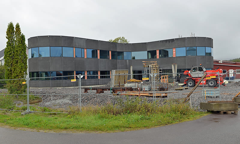 Mørk, buet kontorbygning under bygging, omkranset av gjerder, grus og rød maskin