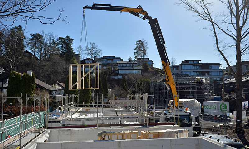 Gul kran heiser trevegg over byggeplass med stillas under blå himmel, trær og moderne hus.
