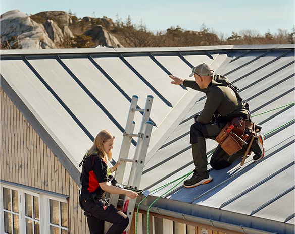 To takarbeidere med verktøybelte og sikkerhetsliner på grått metalltak over lyst trehus.