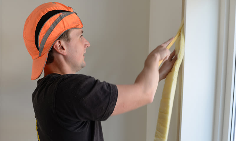 Person i oransje caps monterer gul isolasjonslist i hvit vinduskarm.