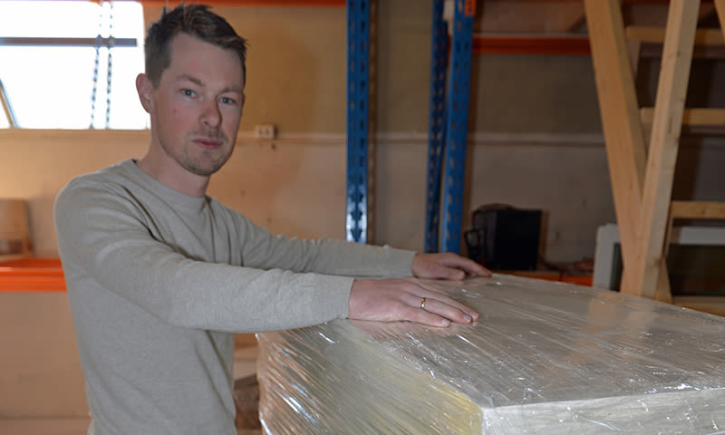 Mann i grå genser støtter hendene på plastpakket pall i lager med blå reoler og trestige.