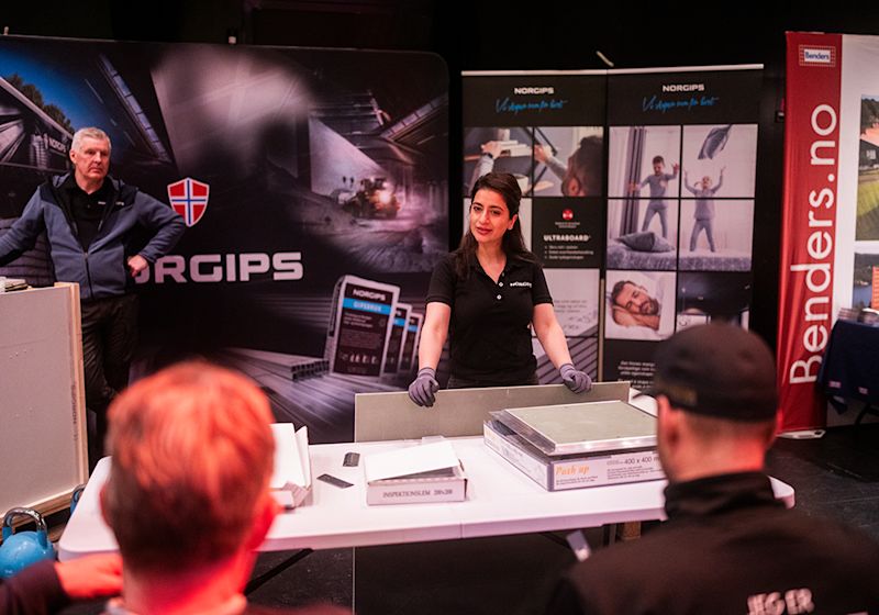 Kvinne i Norgips-uniform presenterer byggematerialer ved en stand på en messe, med publikum i forgrunnen.