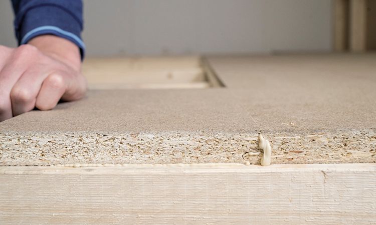 Hånd med blå erme presser sponplate på lys treramme med lim som pipler ut