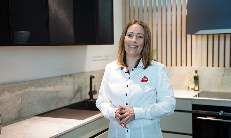 Person i hvit skjorte med rødt BM-merke foran moderne kjøkken med grå benkeplate, svart vask