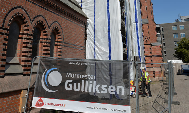 Rød teglkirke under renovering med stillas, banner ’Murmester Gulliksen’ og arbeider i gult