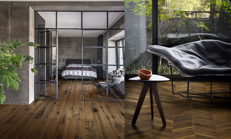 Industrirom med betong, svartrammet glassvegg, varmt treparkett, seng, grønne planter og grå lenestol.